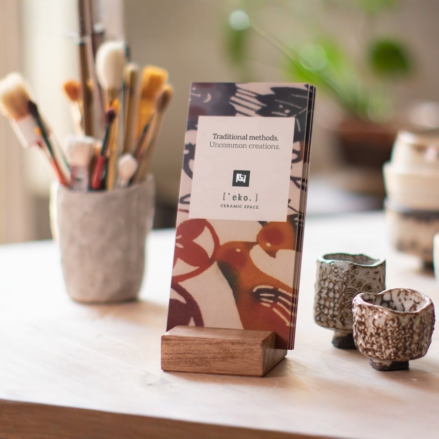 A stack of bi-fold brochures for a ceramics studio displayed on a table in a wooden holder, with ceramic cups and brushes in the background.