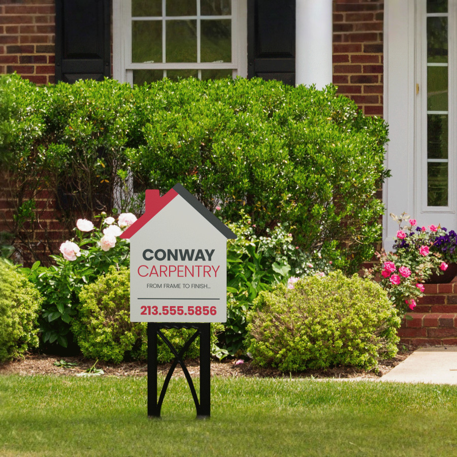 A carpentry yard sign in the shape of a house shown outdoors on a black plastic stand.