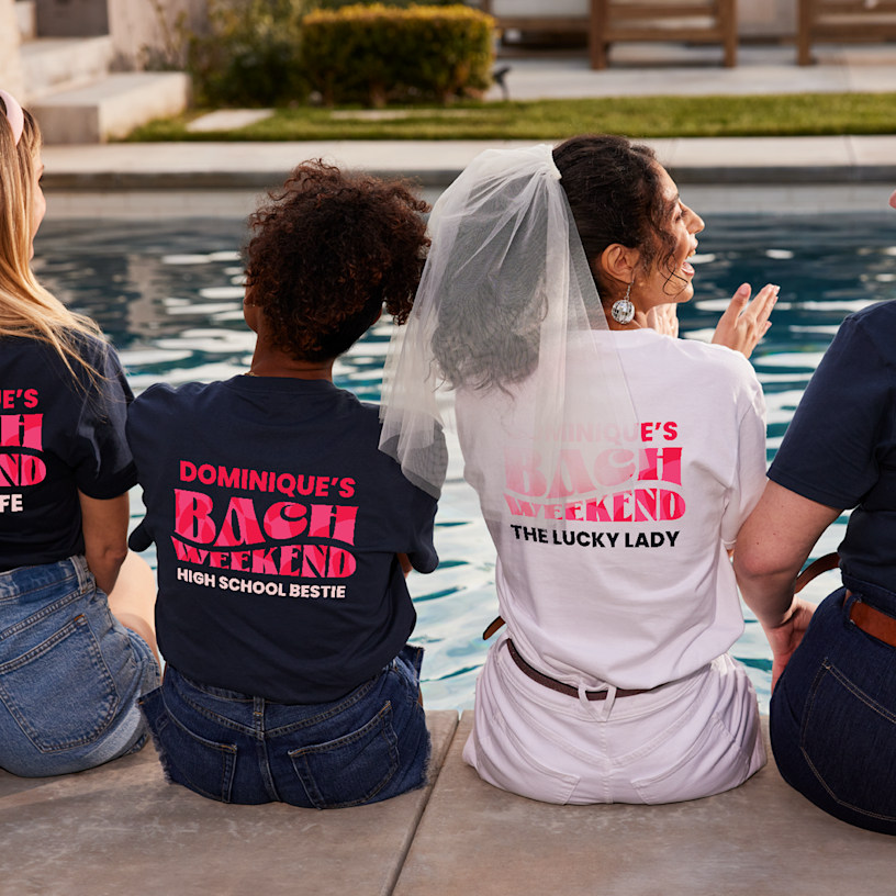 A small group of women sitting by a pool and wearing bachelorette trip shirts.    