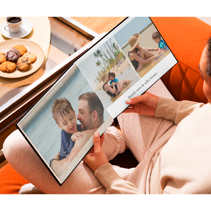 A pair of hands holding an open photo book over a coffee table. The photos show a family at the beach.
