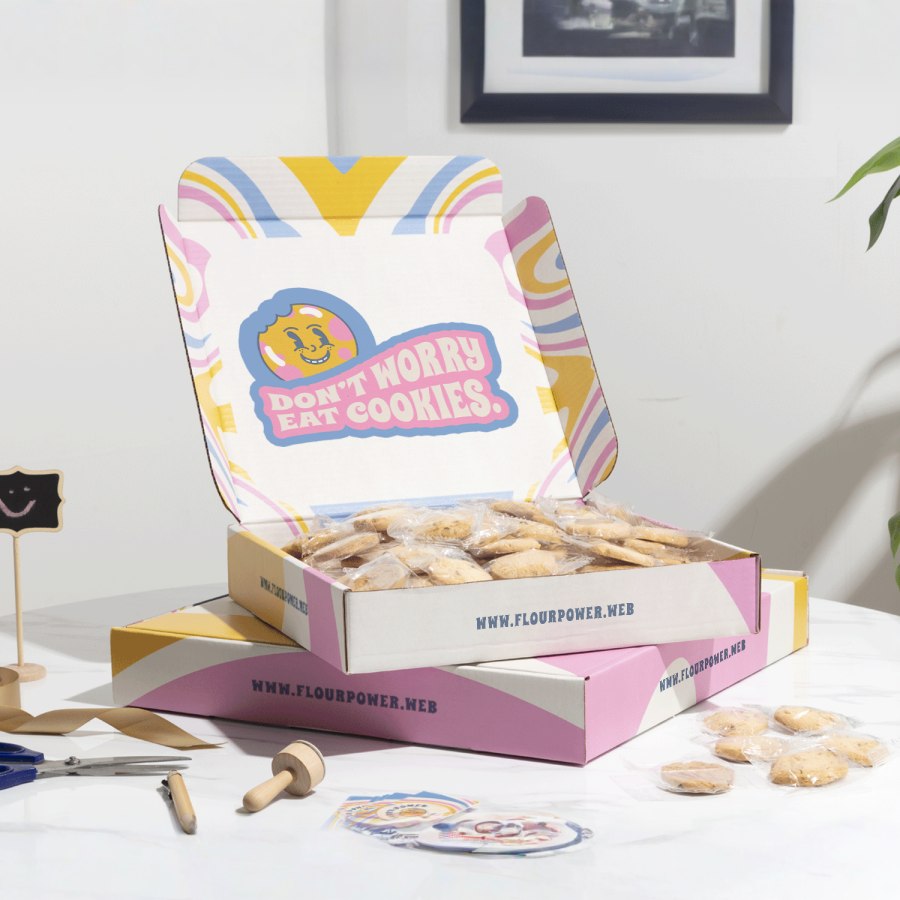 2 square mailer boxes lying flat on top of each other, the top one is open and filled with cookies. The boxes are multi-colored and branded.