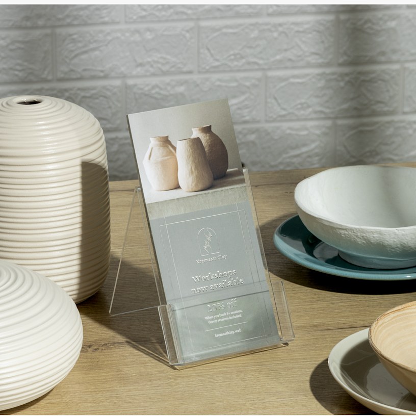 A stack of silver embossed foil rack cards for a ceramics studio. The rack cards are in a plastic holder surrounded by ceramics.