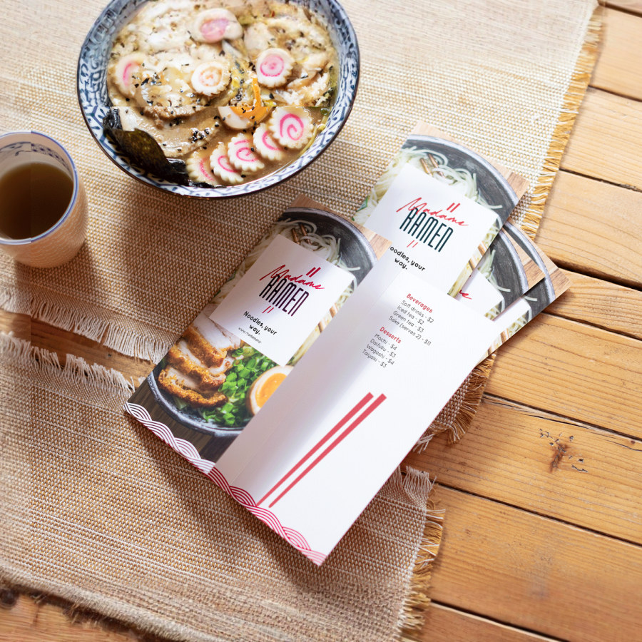 A small stack of tri-fold menus for a ramen restaurant, sitting next to a cup of tea and bowl of soup. 