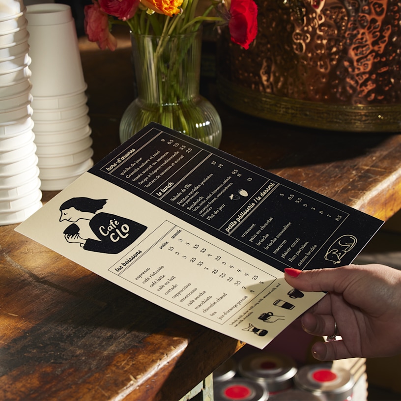 A flat menu for a coffee shop, held by a customer in front of a stack of custom cups. 