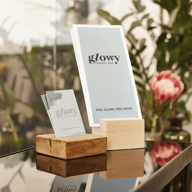 A stack of tri-fold brochures for a beauty brand displayed in a wooden holder, with matching business cards beside them and flowers in the background.