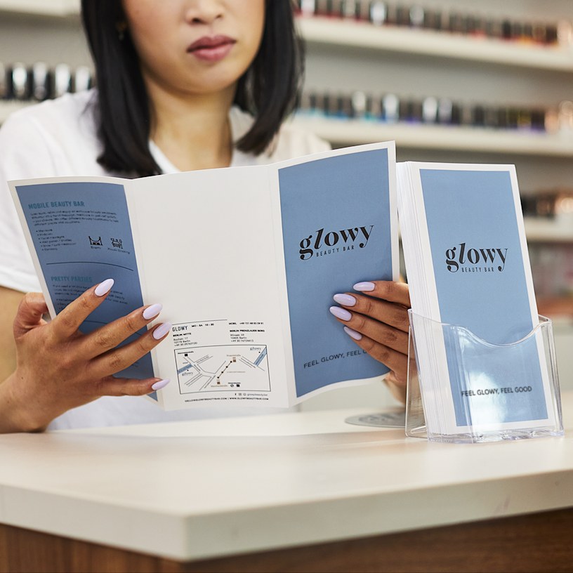 Tri-fold brochures for a beauty brand, displayed in a clear holder on a counter, with someone reading a brochure in the background.