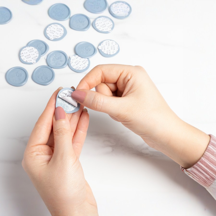 A pair of hands peeling off the backing of a blue custom wax seal. 