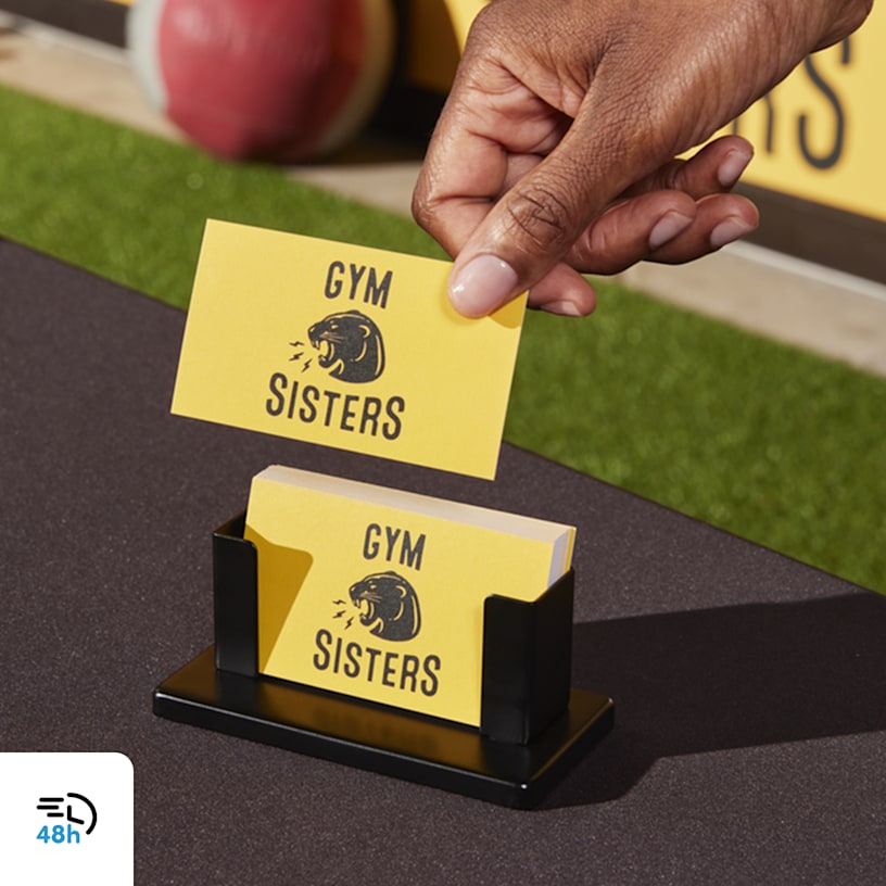 A person picking up a yellow business card for a gym from a small black stand on an outdoor table, with a 48-hour delivery icon in the corner.