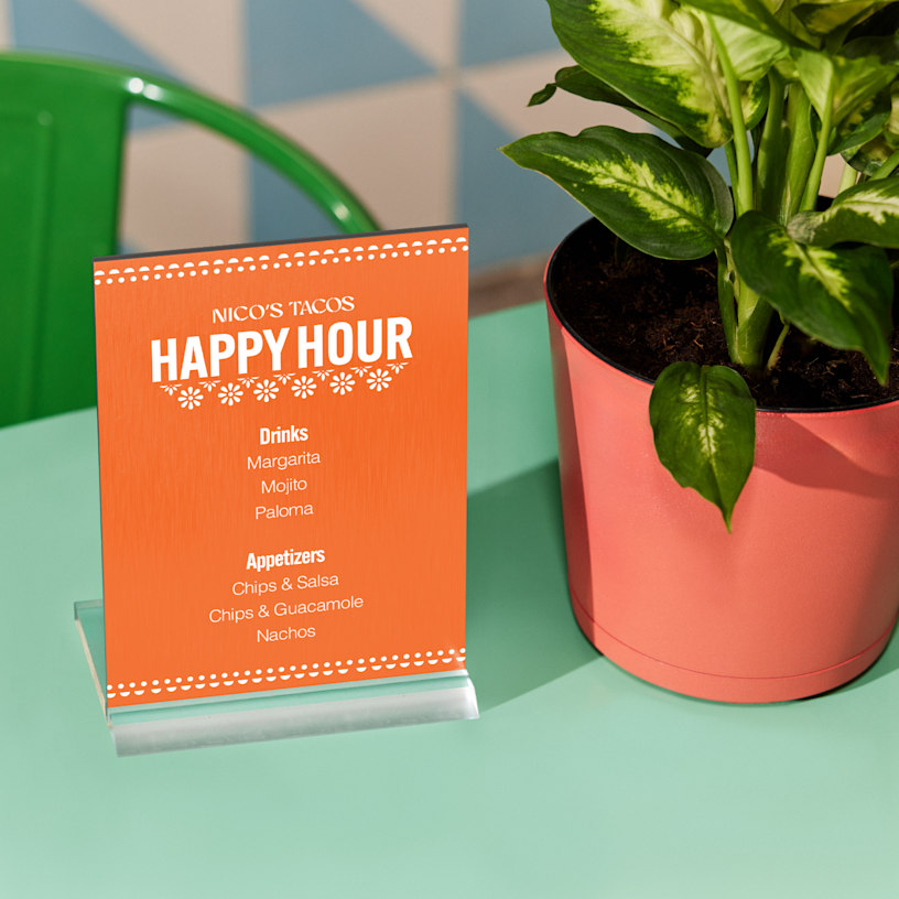 An orange metal table sign showing off happy hour drinks and appetizers for a taco restaurant.