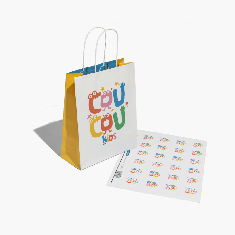 A custom paper bag printed with a kids’ shop logo, set beside a sheet of matching stickers.  