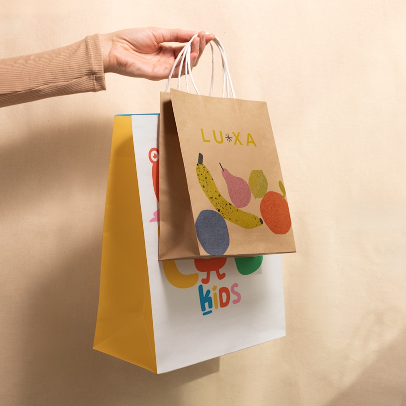 A hand holding up two custom paper bags; one is a kraft bag printed with fruit graphics and the other is a larger white and yellow bag printed with a kids’ shop logo. 