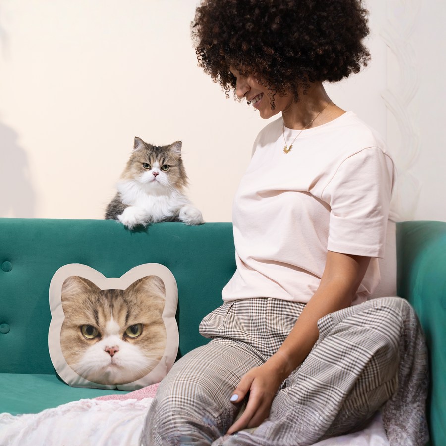 A woman sitting on a couch with her cat while she looks at cat face pillow featuring her furry friend printed on it.