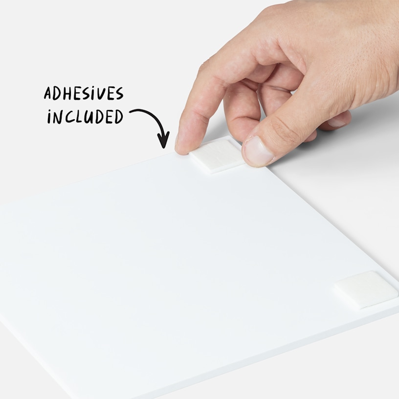 Hand holding a small square piece of double-sided adhesive on the back of a white photo tile.