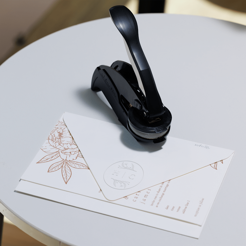 An embossing stamp rests on a white table beside a sealed envelope adorned with floral designs.