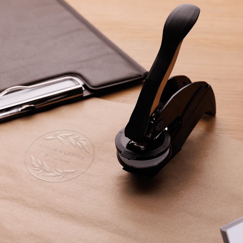 An embossing stamp sits beside kraft personalised stationery, showing a matching, raised impression on the paper.