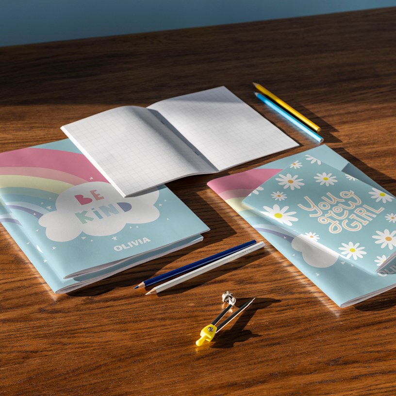 A collection of personalised children's notebooks with colourful designs including unicorn and daisy patterns displayed on a wooden desk with pencils.