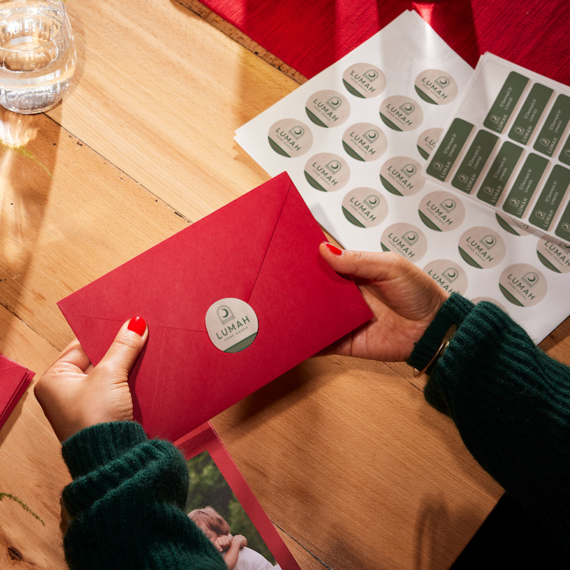 A hand places a round, custom Luxa sticker on a bottle resting on a wooden table, with a sticker sheet next to it.