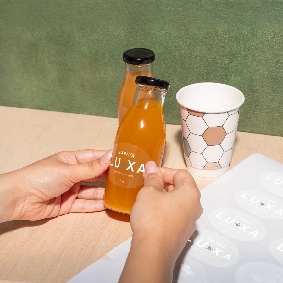 A hand places a round, custom Luxa sticker on a bottle resting on a wooden table, with a sticker sheet next to it.