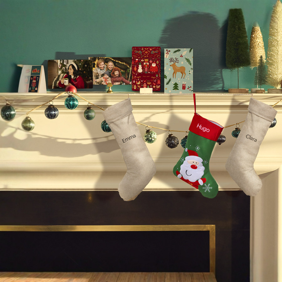 Three personalised Christmas stockings with names hanging on a fireplace mantel, including a Santa stocking and two natural burlap styles.