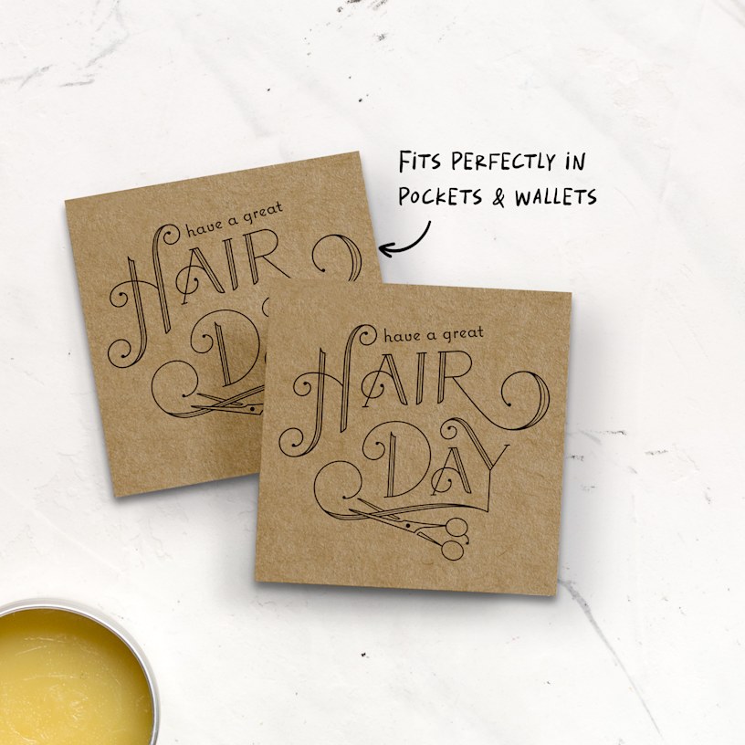 Two square kraft business cards featuring a hairdresser's business are displayed on a counter. The scene includes a pair of scissors and hair Vaseline.