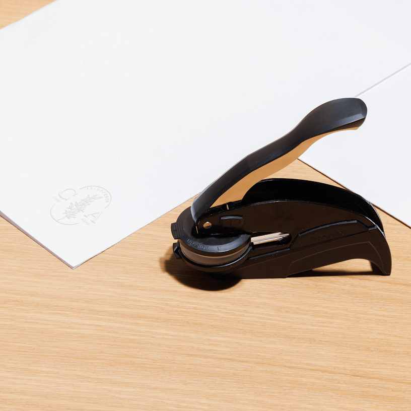 An embossing stamp sits beside white, personalised stationery, showing a matching raised impression on the paper.