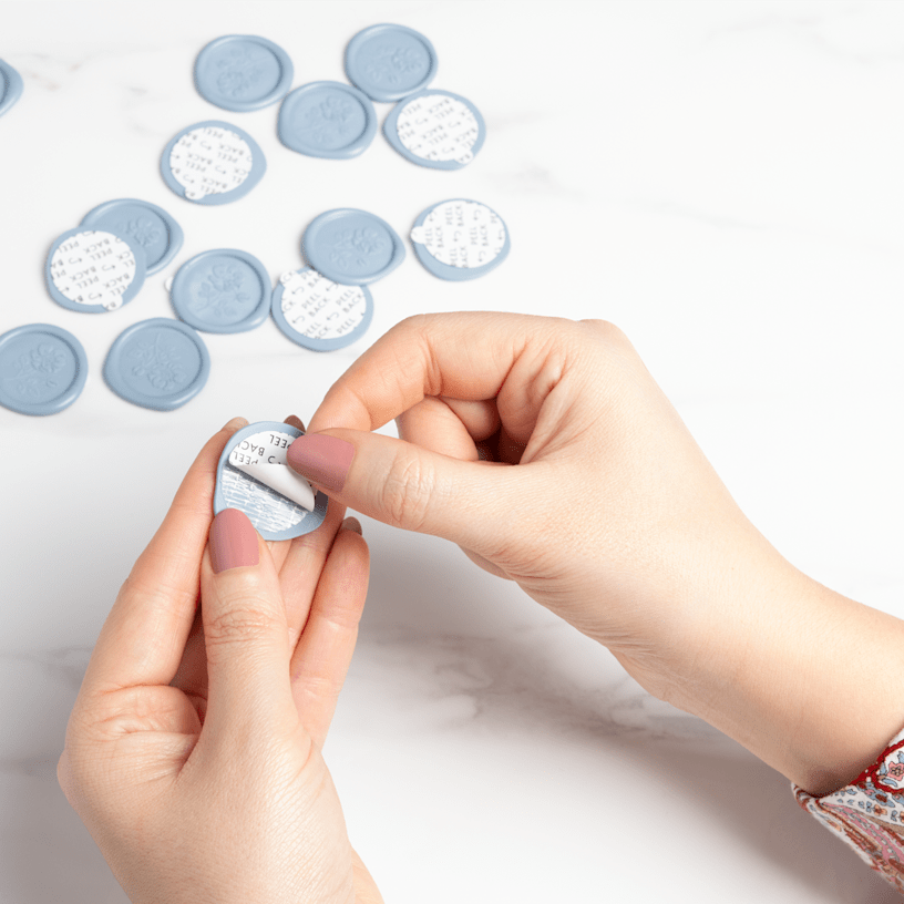 A pair of hands peeling off the backing of a blue, custom wax seal. 