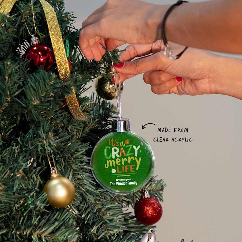 A person hanging up a Christmas bauble featuring a festive quote. An arrow points to the clear acrylic material.