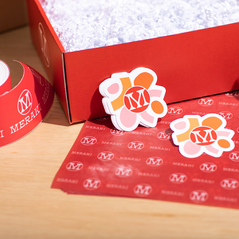 Two custom die-cut stickers shaped like a brand logo, displayed alongside branded roll labels, a mailer box and tissue paper.