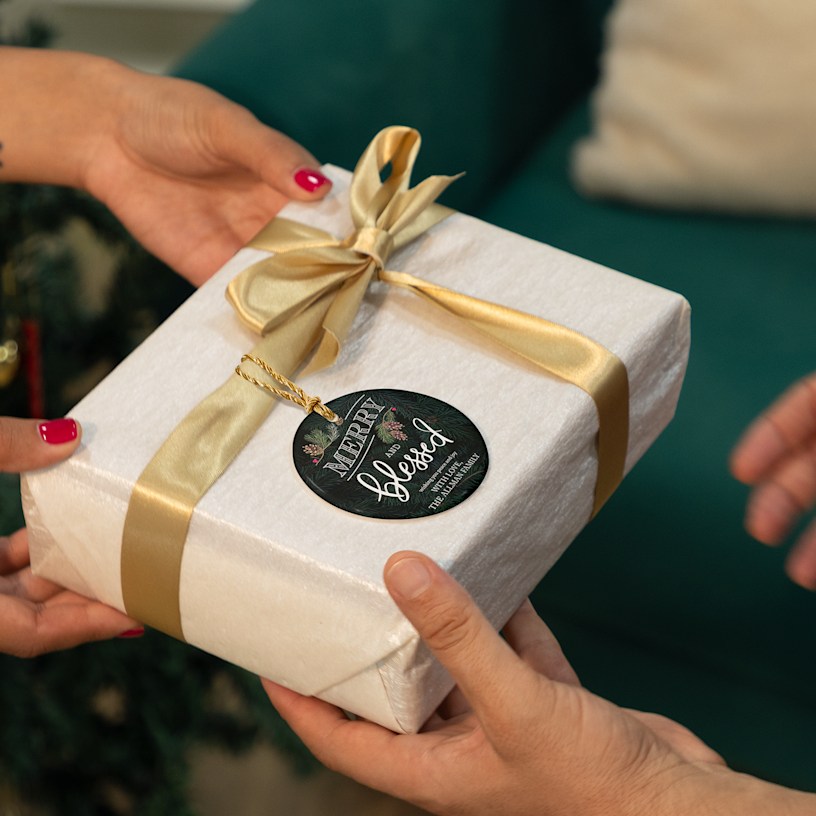 A person handing a wrapped present with a Christmas ornament looped around the ribbon. The ornament features a stylised Christmas greeting.