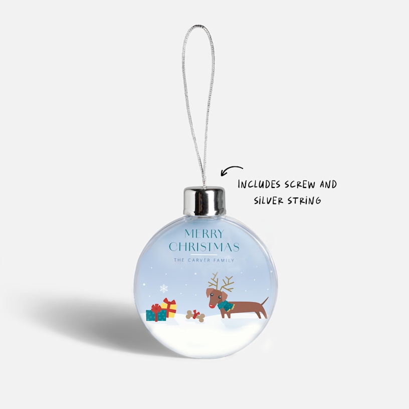 A Christmas bauble featuring an illustrated reindeer and text. An arrow points to the included screw and silver string.