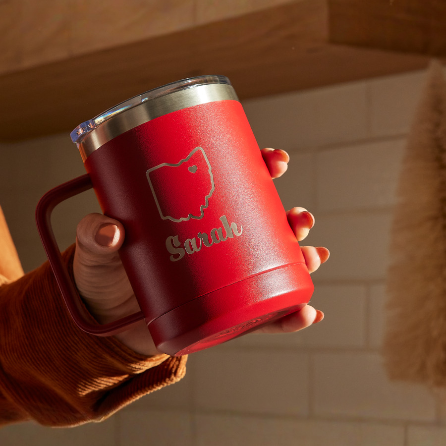 A hand holding a 15 oz. red travel mug at an angle. The mug is laser etched with the name Sarah and the outline of a US state. 