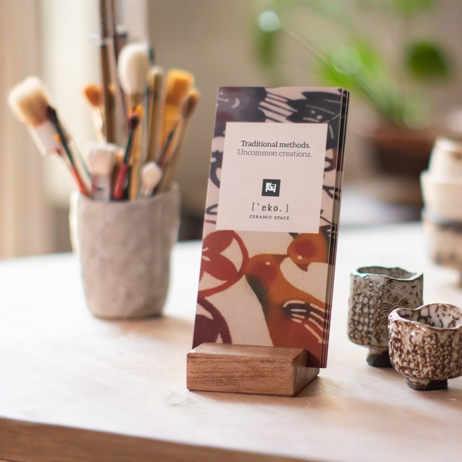 A stack of bi-fold brochures for a ceramics studio displayed on a table in a wooden holder, with ceramic cups and brushes in the background.