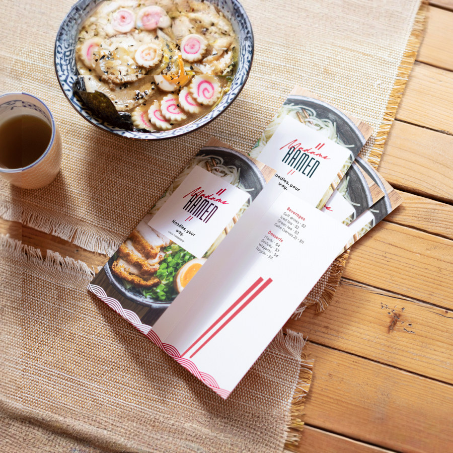 A small stack of tri-fold menus for a ramen restaurant, sitting next to a cup of tea and bowl of soup. 
