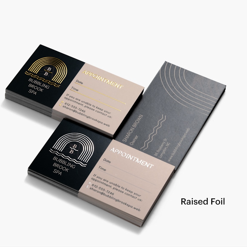 A stack of gold raised foil appointment cards and a stack of silver raised foil appointment cards, both for the same spa and using the same design.