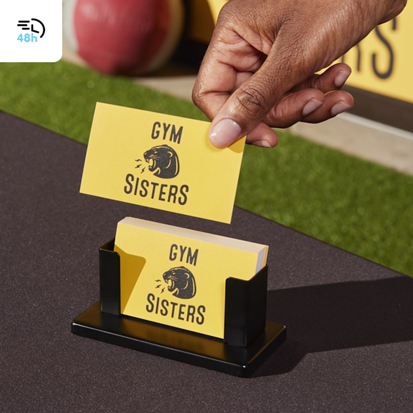 A person picking up a yellow business card for a gym from a small black stand on an outdoor table, with a 48-hour delivery icon in the corner.