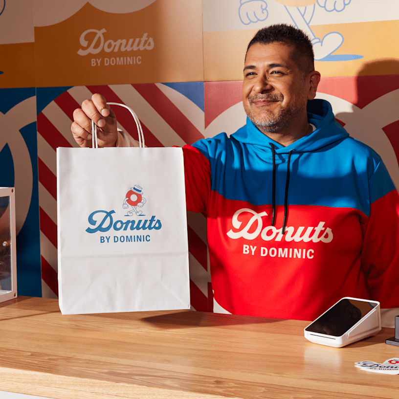A smiling donut shop employee handing a branded kraft paper bag to a customer.