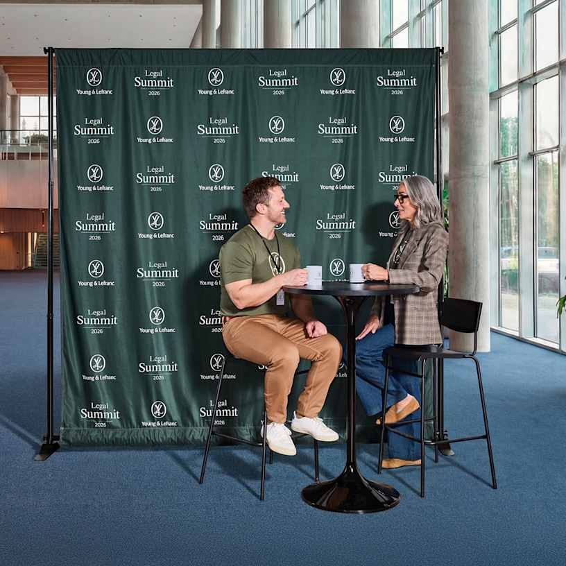 A man and a woman having coffee in front of a step & repeat banner with a logo and business name printed all over it.