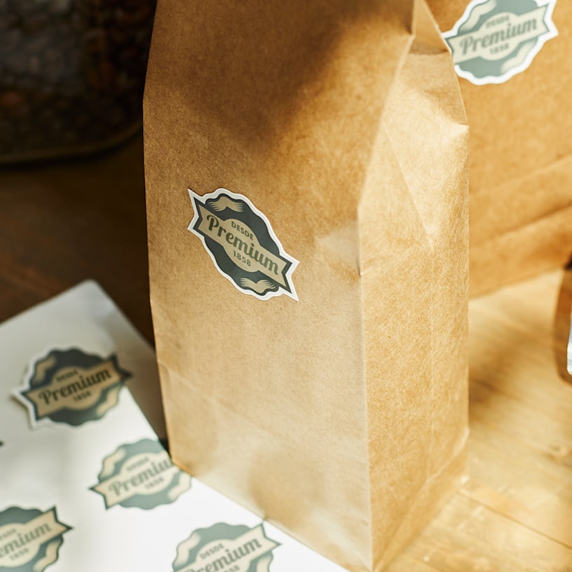 Una bolsa de papel marrón con una etiqueta que dice «Premium» sobre una superficie de madera junto a una hoja de pegatinas personalizadas.