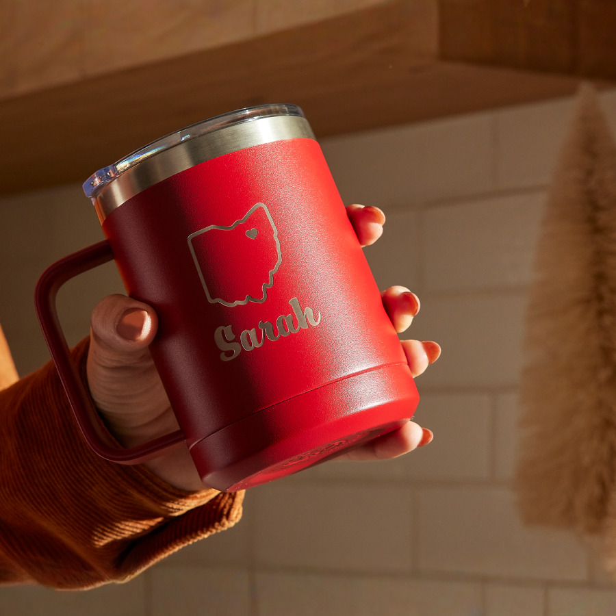 Una mano sosteniendo una taza de viaje roja de 15 oz a un ángulo. La taza está grabada a láser con el nombre Sarah y la de un estado de Estados Unidos. 