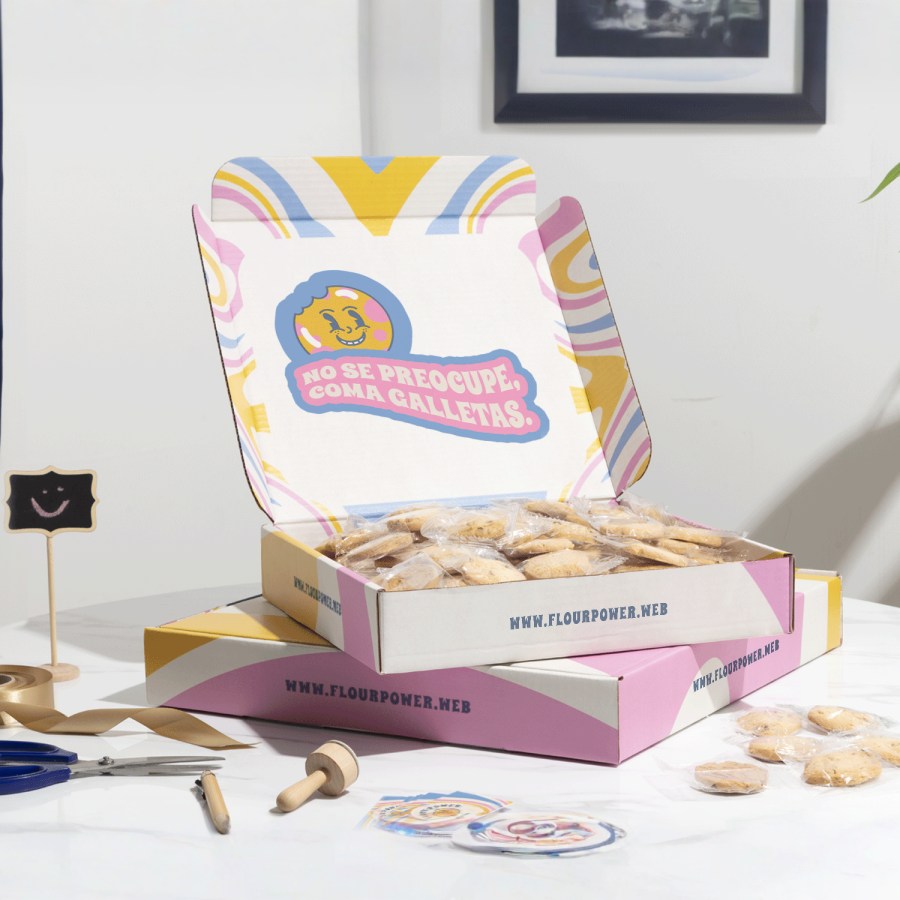 2 cajas postales cuadradas colocadas de manera plana una sobre otra, la de arriba está abierta y llena de galletas. Las cajas son multicolor y con marca.
