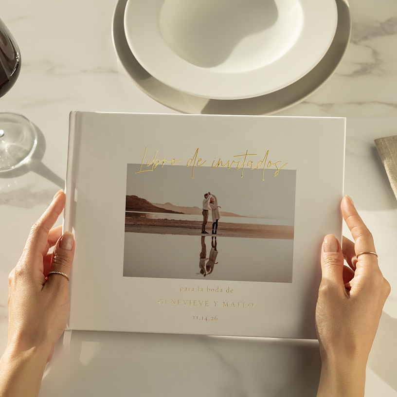 Un par de manos que sostienen un libro de invitados en metalizado dorado, con una elegante mesa puesta al fondo. 