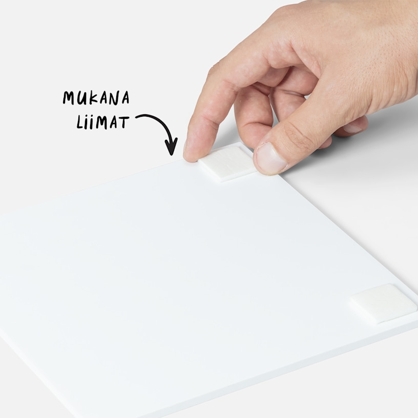 Käsi, joka kiinnittää kaksipuolista teippiliuskaa valokuvataulun kääntöpuolelle, ja maininta siitä, että teippi toimitetaan taulujen mukana. 