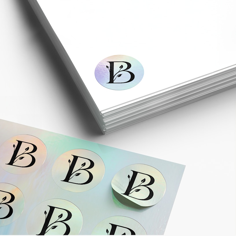 Une planche de stickers personnalisés à côté d’une pile de papiers blancs. Il y a un sticker holographique rond avec la lettre B et un graphisme en forme de feuille sur les papiers.