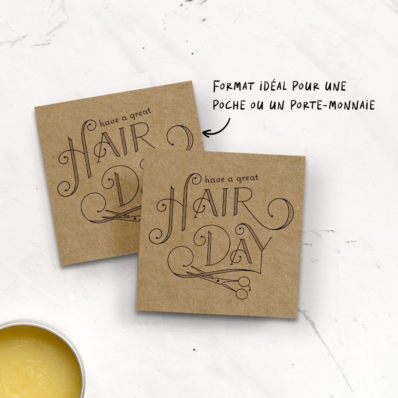 Deux cartes de visite carrées en papier kraft pour un salon de coiffure sont placées sur un comptoir. La scène inclut une paire de ciseaux et de la vaseline pour cheveux.
