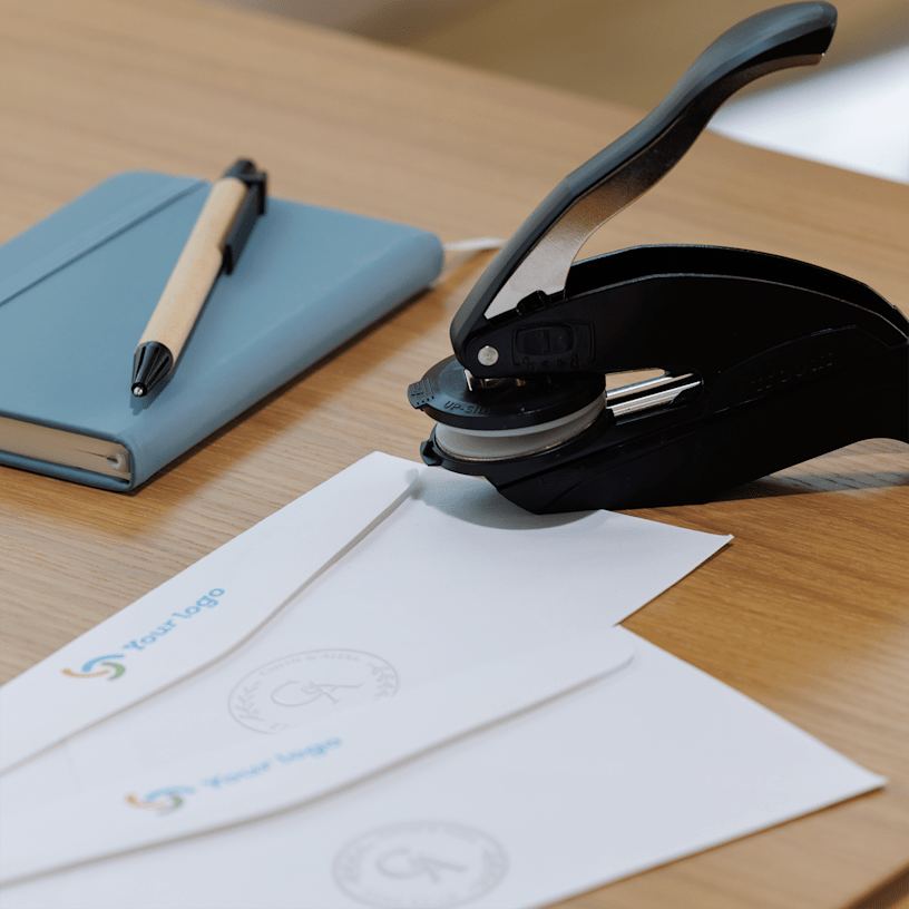 Un bloc-notes, un stylo et une pince à gaufrer sont posés sur un bureau, à côté de deux enveloppes blanches, chacune marquée d’une impression ronde gaufrée.
