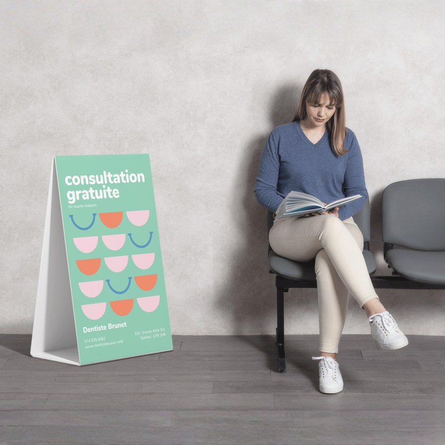 Image d'une femme en chemise bleue assise dans une salle d’attente en train de lire, avec un panneau sur pieds en carton ondulé faisant la promotion de consultations gratuites avec un dentiste.