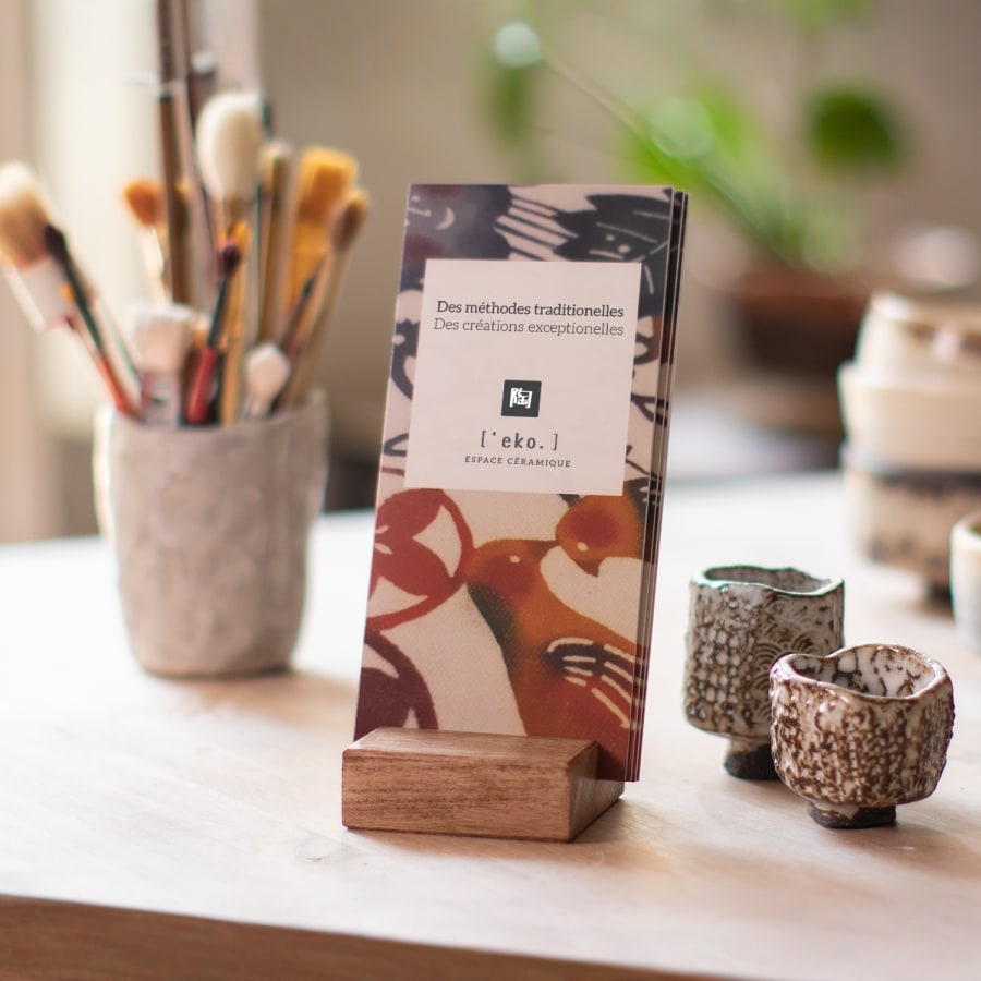 Une pile de dépliants à deux volets créés pour un atelier de céramique sont présentés dans un porte-dépliants en bois posé sur une table, avec des tasses en céramique et des pinceaux en arrière-plan.