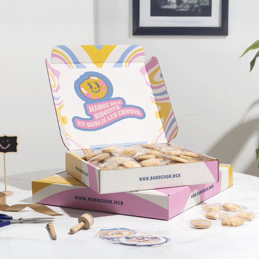Deux boîtes carrées pour envoi postal superposées, dont celle du dessus, ouverte, est remplie de biscuits. Les boîtes sont multicolores et personnalisées.