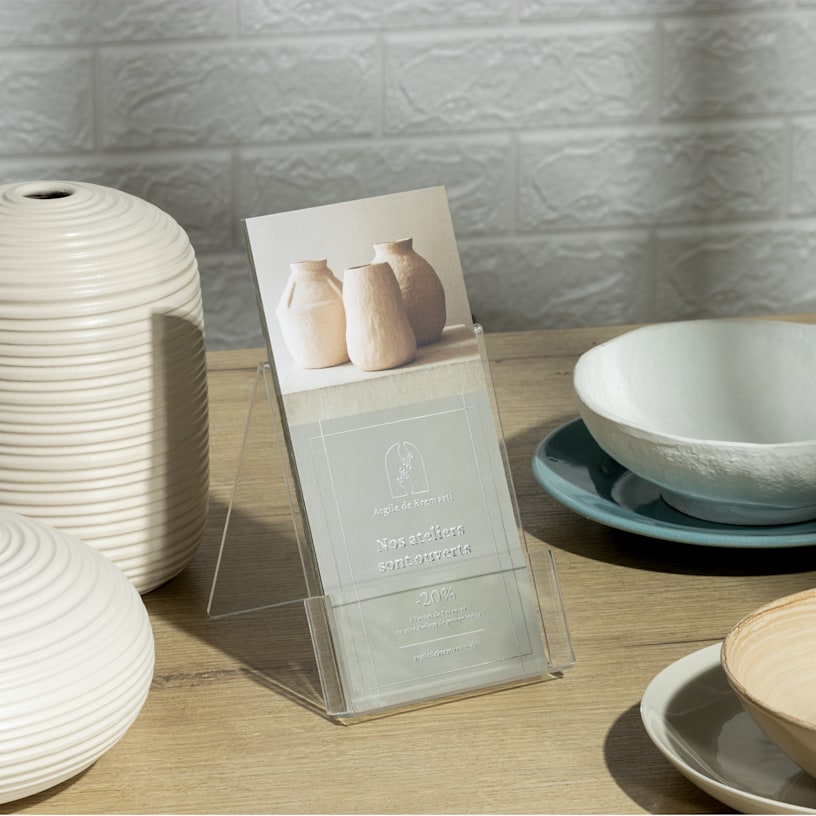 Pile de cartons d’information avec effet métallisé argenté en relief faisant la promotion d’un atelier de céramique. Les cartons d’information sont rassemblés dans un porte-cartes en plastique entouré de poteries en céramique.