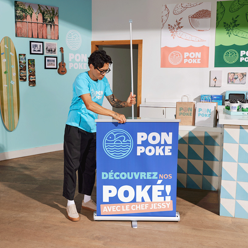 Au cœur d’un restaurant animé, un homme installe une banderole rétractable pour présenter ses bols de poké.